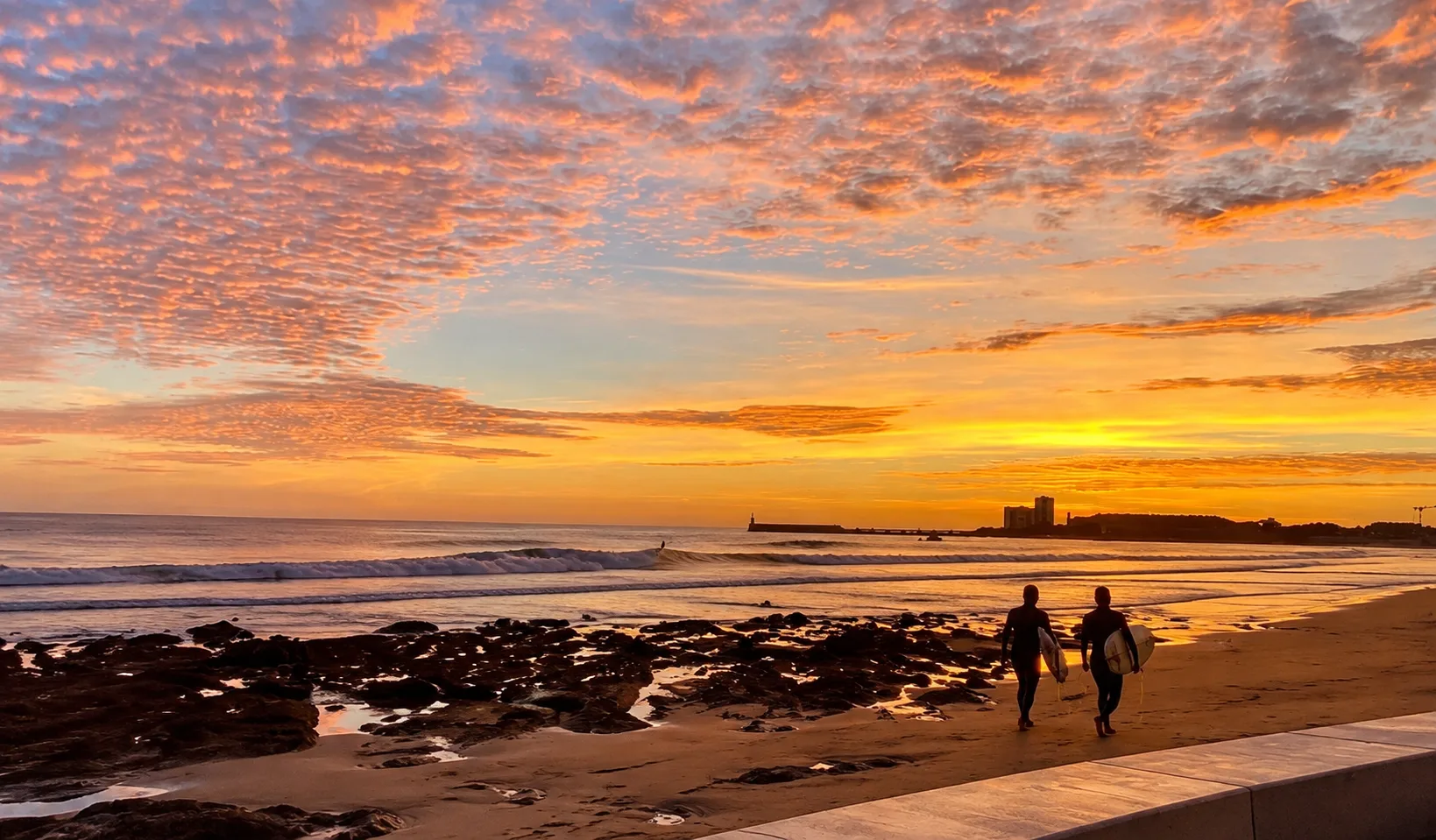 Deux surfeurs marchent sur le sable au coucher du soleil, planche sous le bras, la jetée et le phare des Sables-d’Olonne au fond.