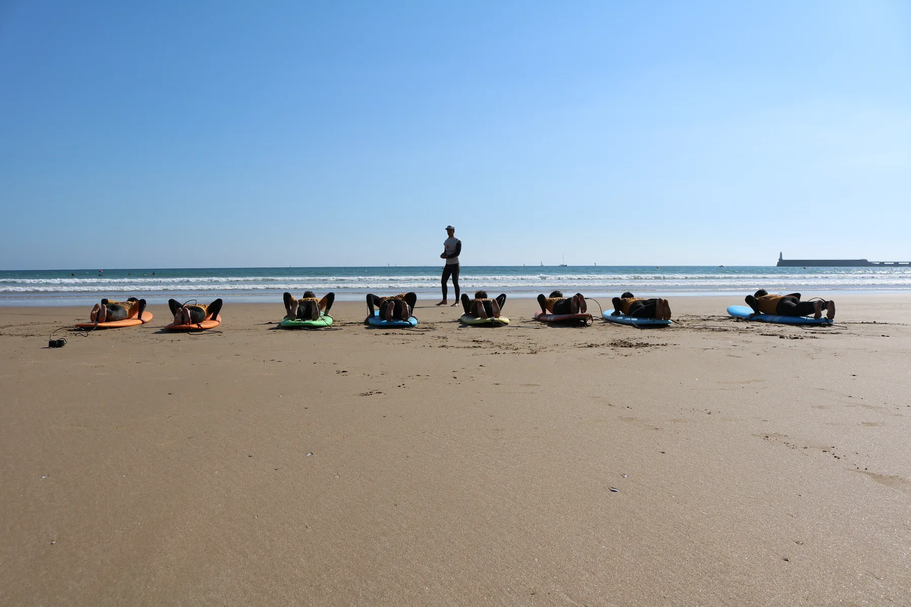 Huit élèves allongés sur leurs planches sur le sable, en arc de cercle devant le moniteur.